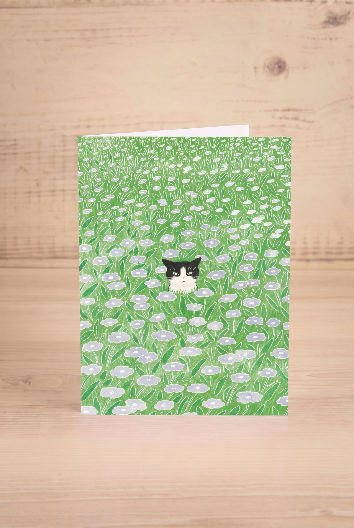 A vertical greeting card featuring a watercolor-style green field of flowers with a small tuxedo cat by Foonie at maison garçonne in montreal