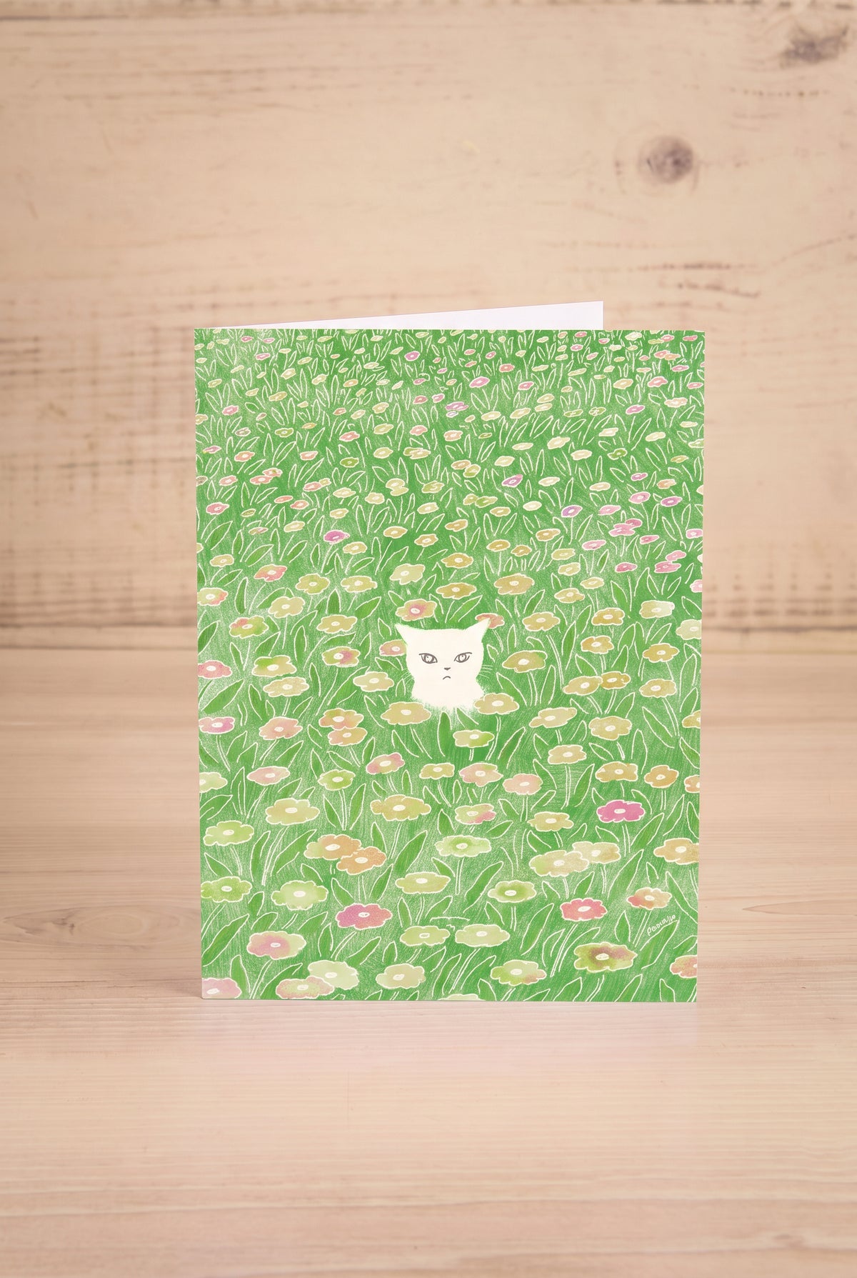 A vertical greeting card featuring a watercolor-style green field of flowers with a small white cat by Foonie at maison garçonne in montreal