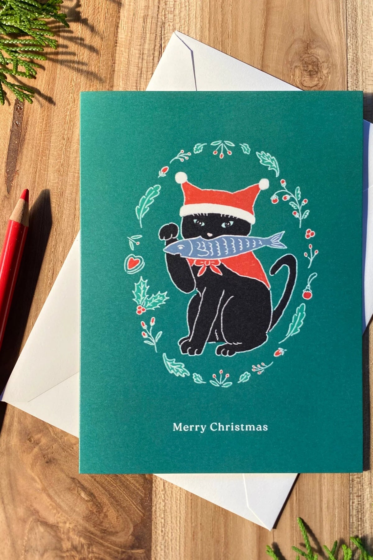 A Christmas greeting card featuring a black cat wearing a Santa hat and holding a silver fish, surrounded by a wreath, placed on top of a white envelope on a wooden surface at la maison garçonne in montreal.