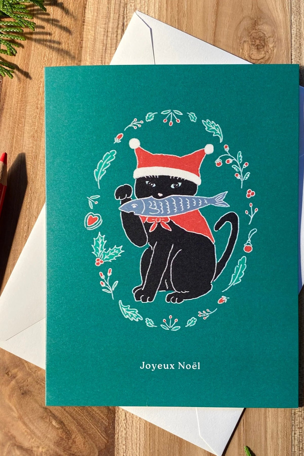A teal Christmas greeting card with an illustration of a black cat wearing a Santa hat and scarf while holding a silver fish, placed on top of a white envelope on a wooden surface at la maison garçonne in montreal.