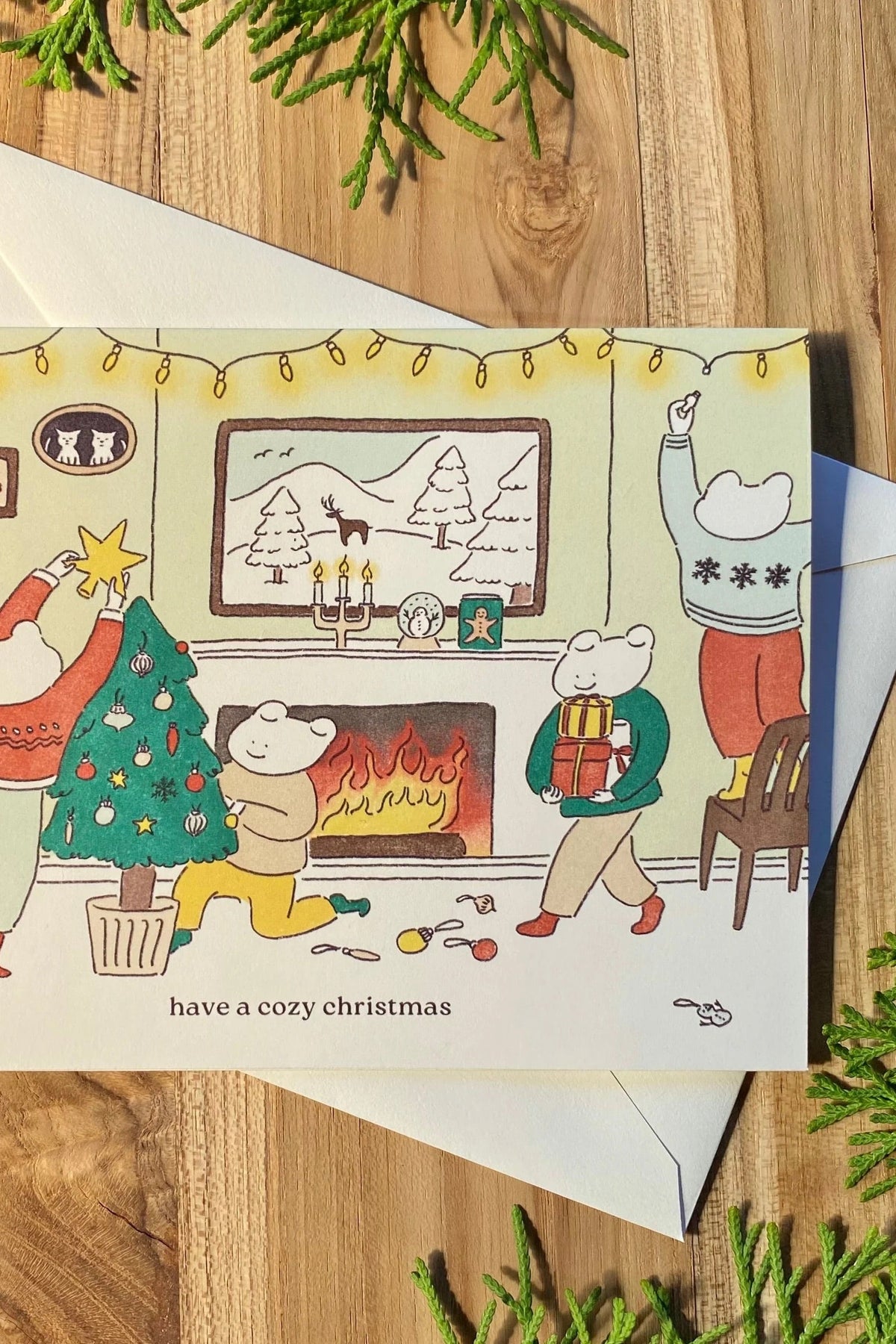 A Christmas greeting card by Foonie with a cozy illustrated scene of people decorating a tree and gathering around a fireplace, shown with a white envelope on a wooden surface at la maison garçonne in montreal