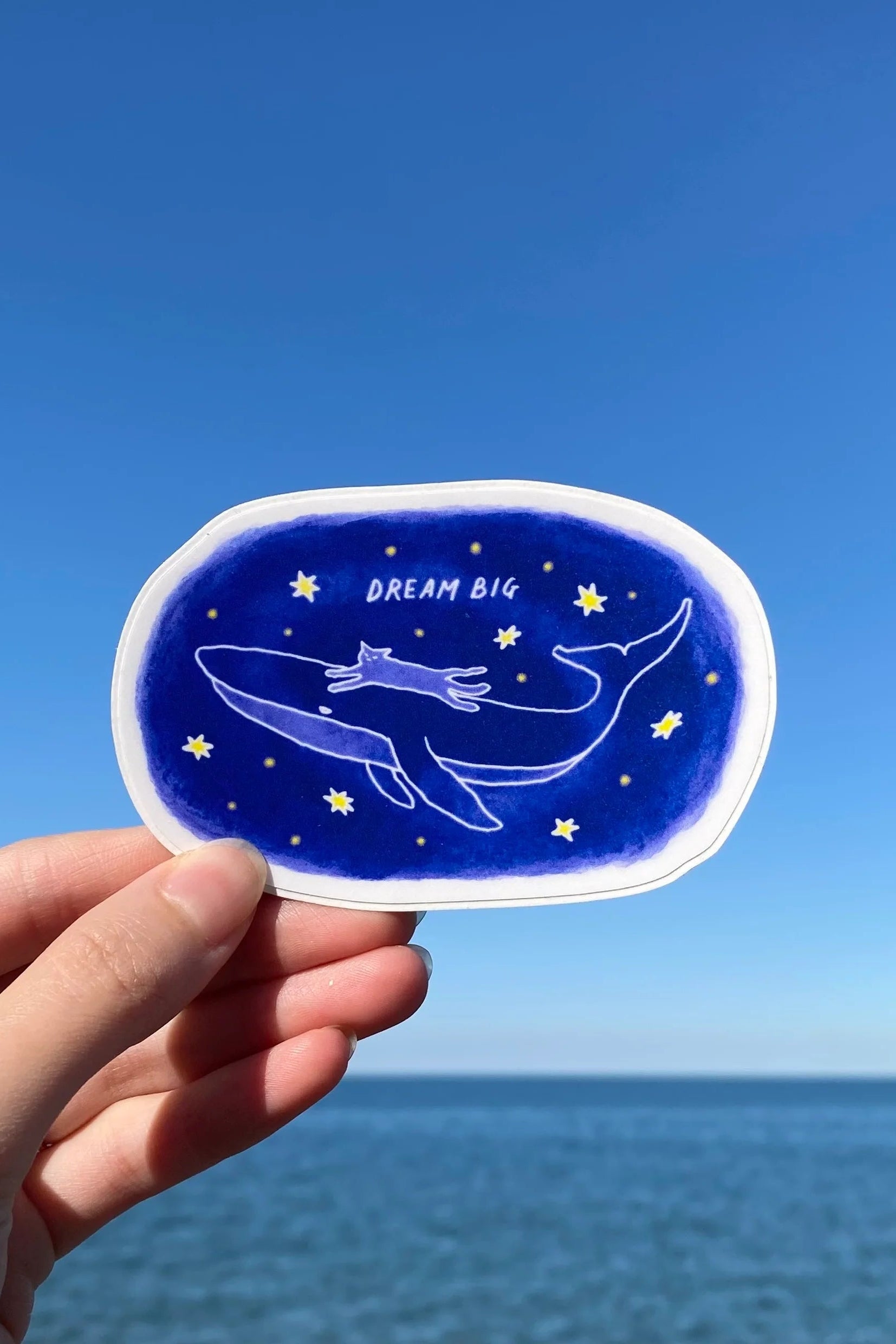 Hand holding a blue sticker from Foonie with a whale design and 'Dream Big' text against a clear blue sky.
