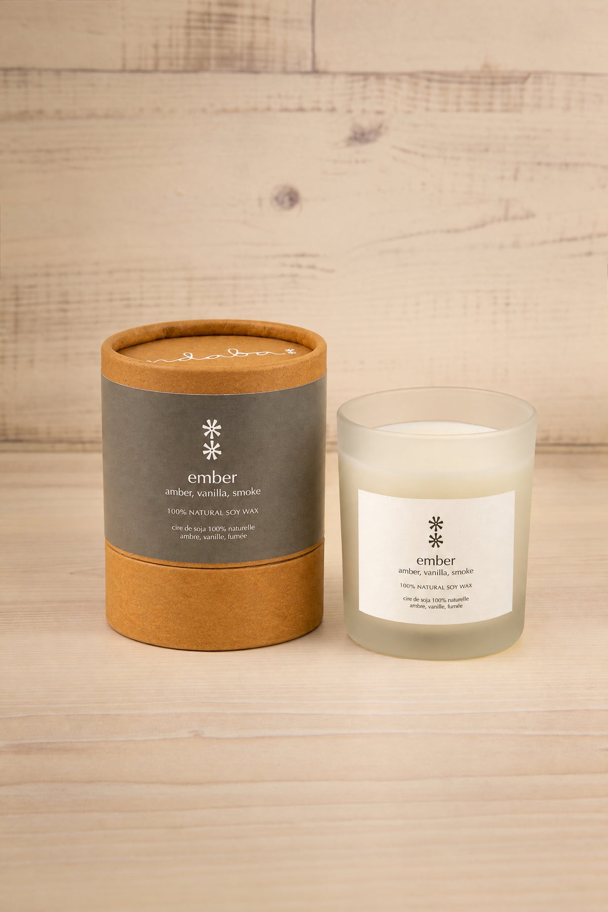 A candle product shown with its cylindrical kraft paper packaging and the glass jar candle beside it on a light wood surface against a light wood plank background at la maison garçonne. The labels read 'ember' with a snowflake icon.