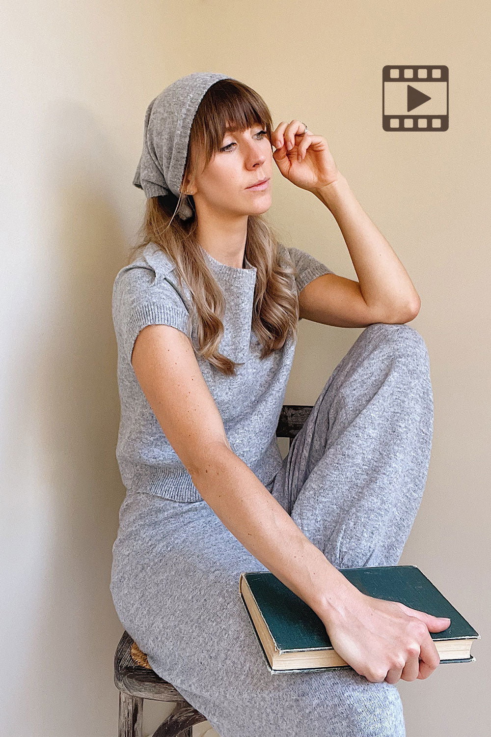 Photo of a mannequin wearing the Kano Grey outfit sitting on a stool with a book, against a plain wall.