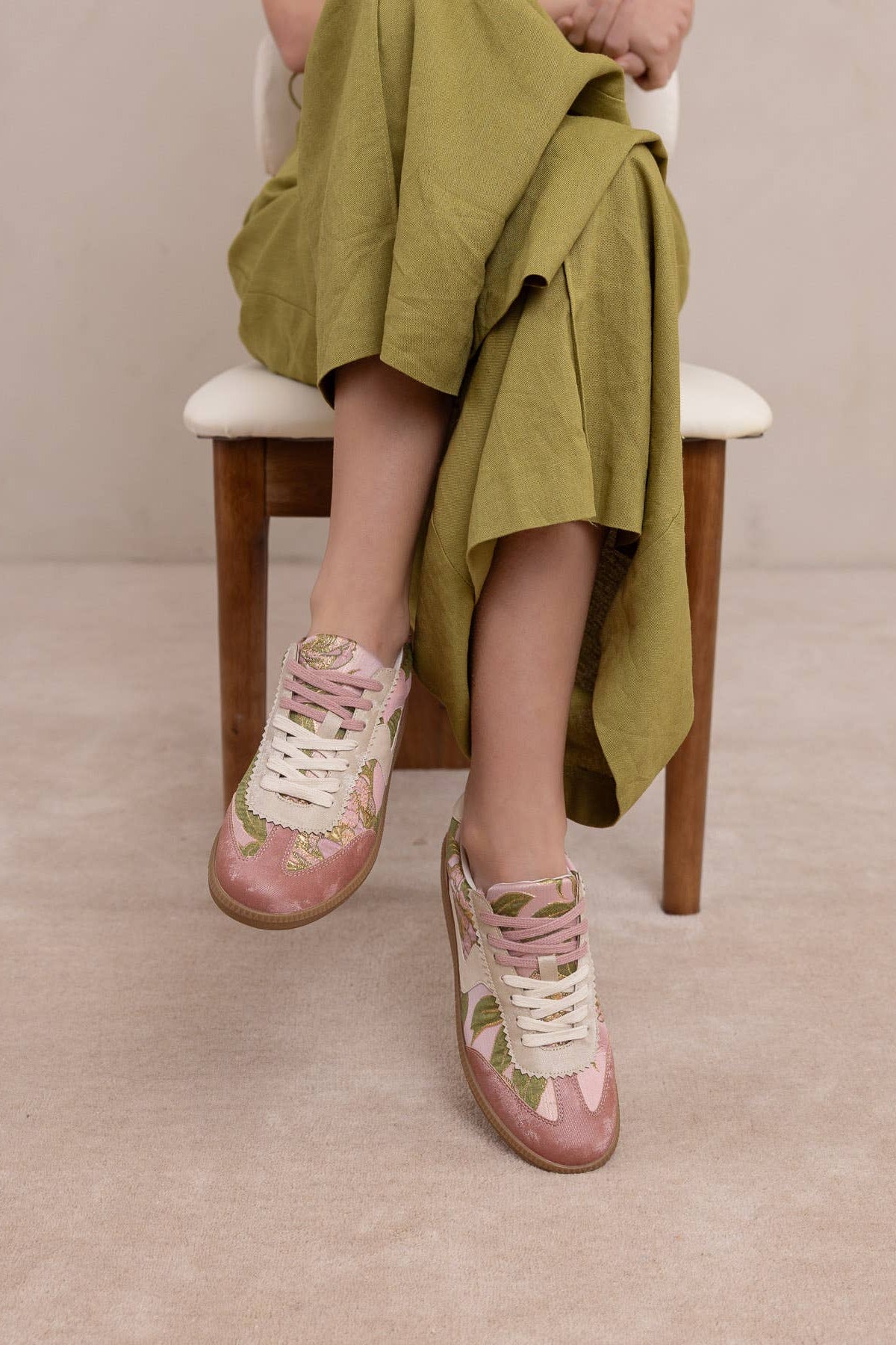 Woman wearing a pair of low-top sneakers in dusty pink suede and glossy pink patent leather, decorated with a green floral print, cream laces, and gum soles, displayed on a textured pink background at boutique 1861.