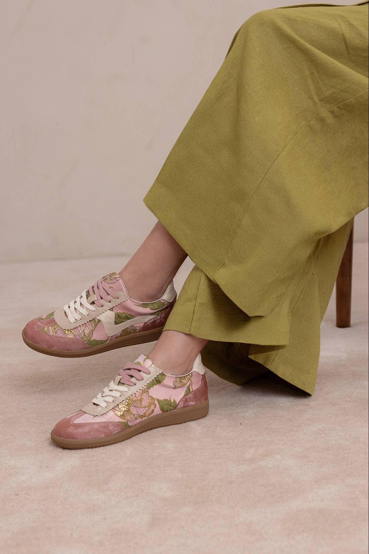 Woman wearing a pair of low-top sneakers in dusty pink suede and glossy pink patent leather, decorated with a green floral print, cream laces, and gum soles, displayed on a textured pink background at boutique 1861.