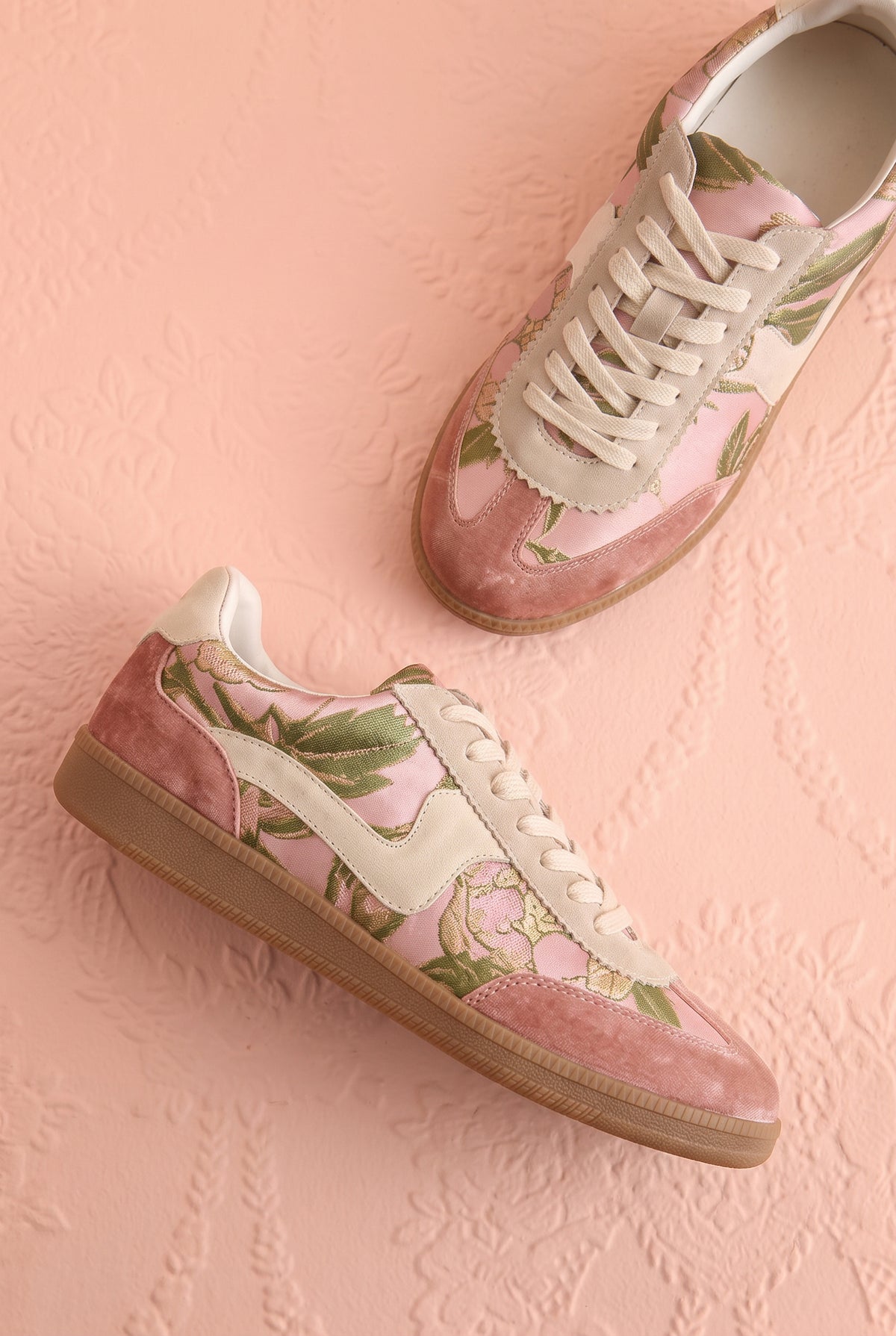 Top view of a pair of low-top sneakers in dusty pink suede and glossy pink patent leather, decorated with a green floral print, cream laces, and gum soles, displayed on a textured pink background at boutique 1861.