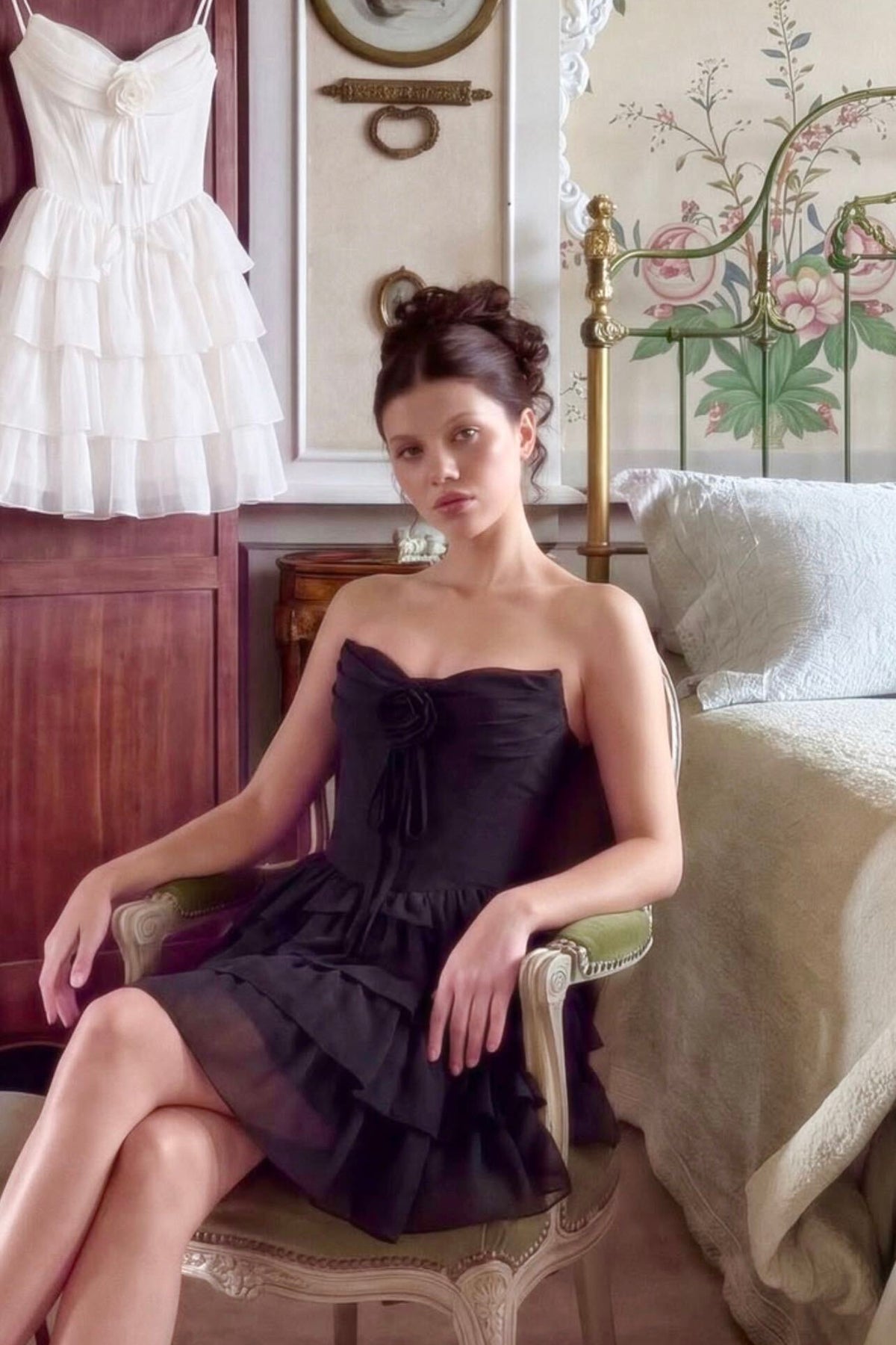 A woman sits on an ornate chair in a bedroom setting, wearing Naemia Black Strapless Corset Mini Dress with Flower from boutique 1861 montreal