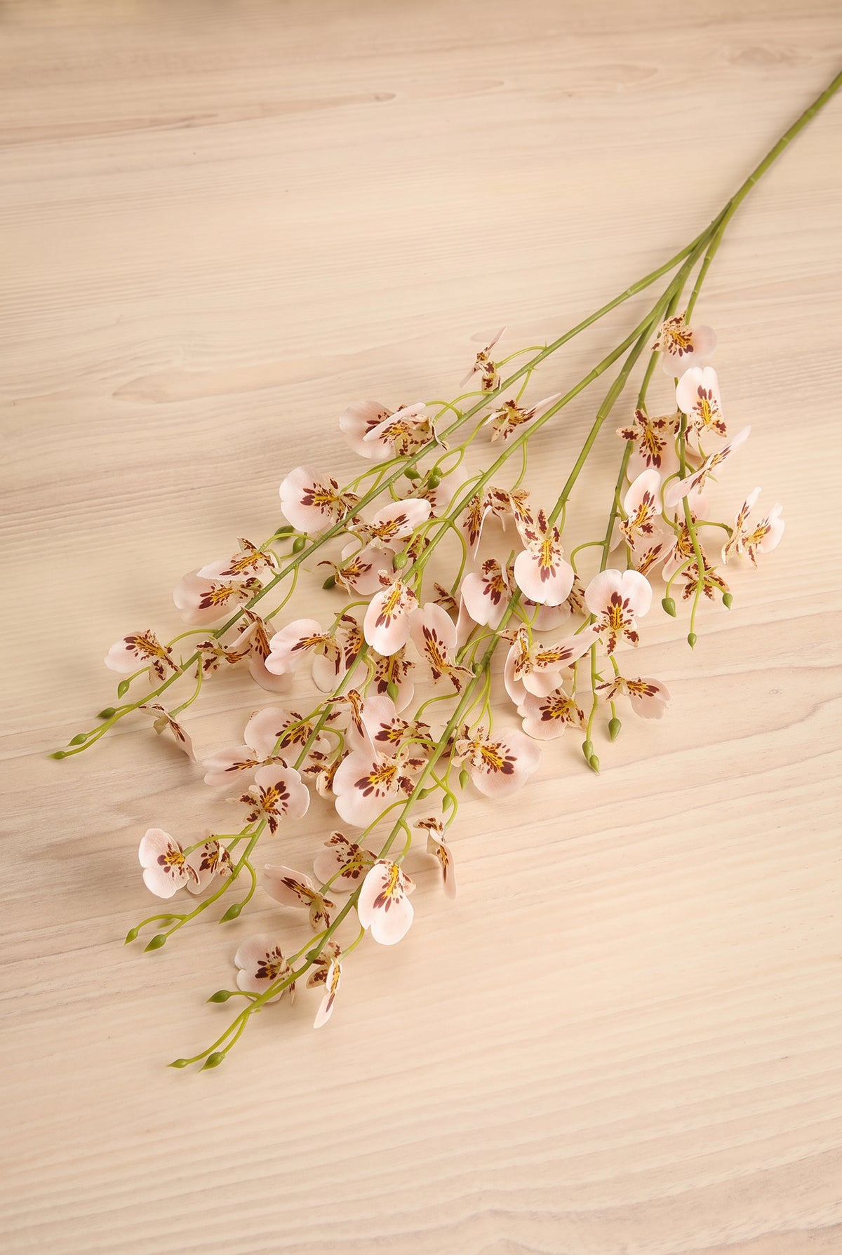 A bouquet of artificial light pink wild orchid spray flowers with green stems, arranged diagonally on a light beige wood-grain surface at la maison garçonne in montreal.