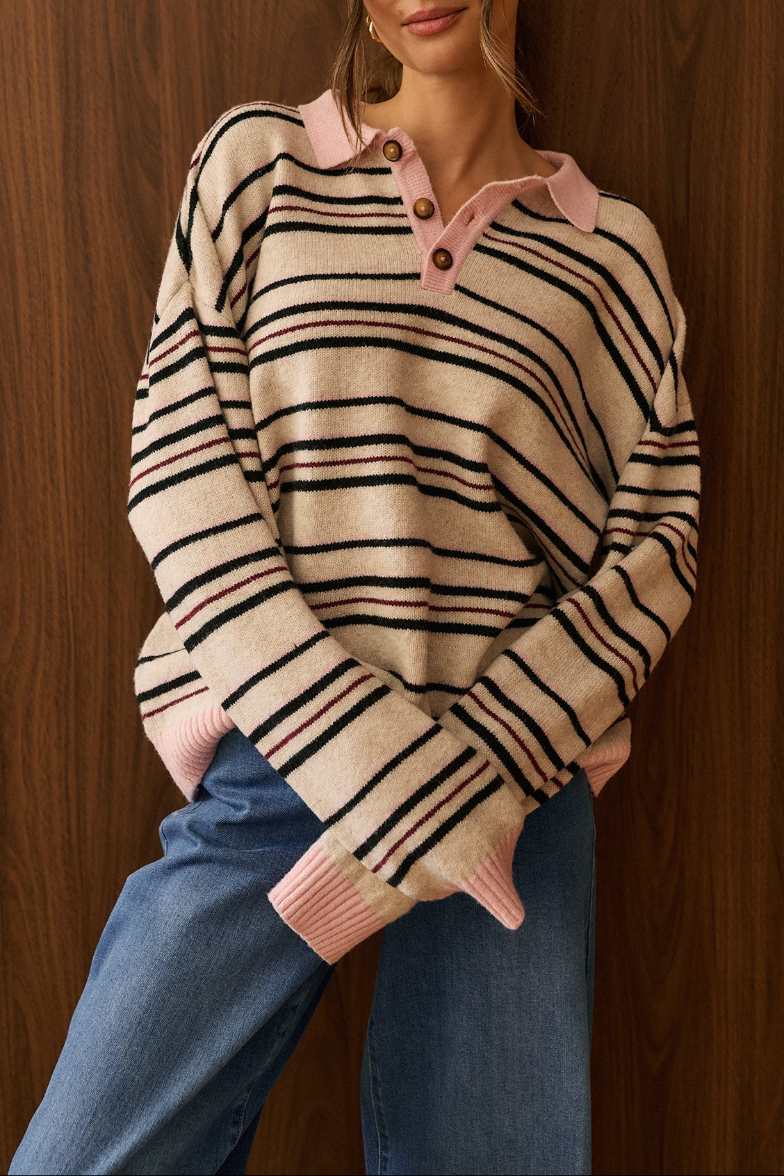 A woman stands against a dark wood-paneled wall wearing a beige knit sweater Gabbie with thin black horizontal stripes and a pink collar by dailystory Montreal leaning against a dark wood-paneled wall.