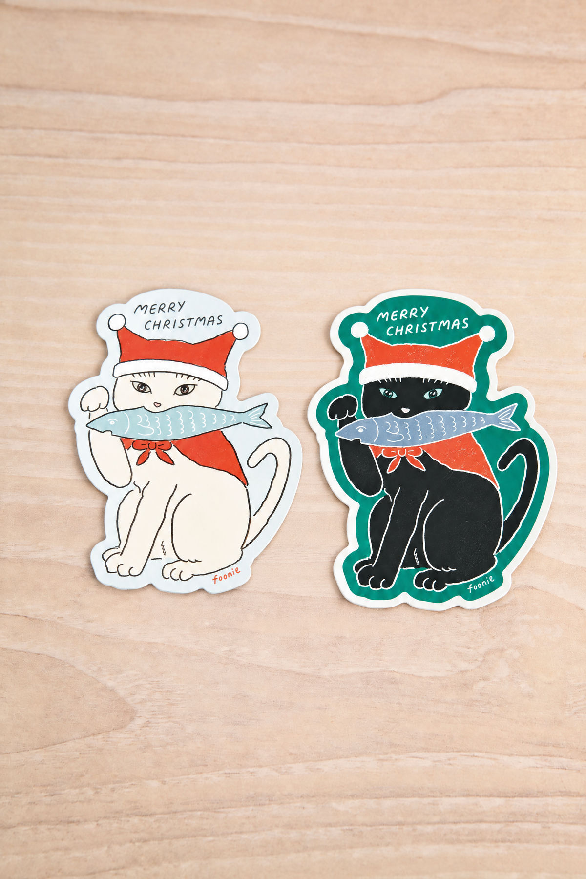 Two Christmas-themed cat stickers from Foonie on a light wood background at maison garçonne in montreal. One sticker shows a white cat wearing a red Santa hat and scarf while holding a blue fish, with the text 'MERRY CHRISTMAS'.