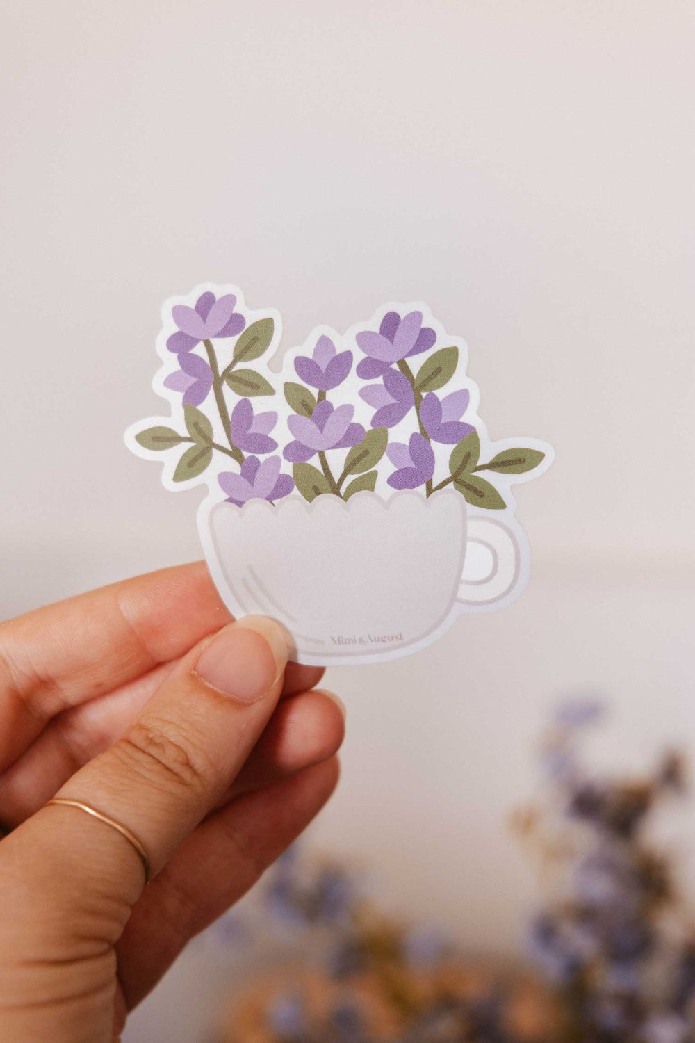 Hand holding a sticker of purple flowers in a white cup from mimi and august 