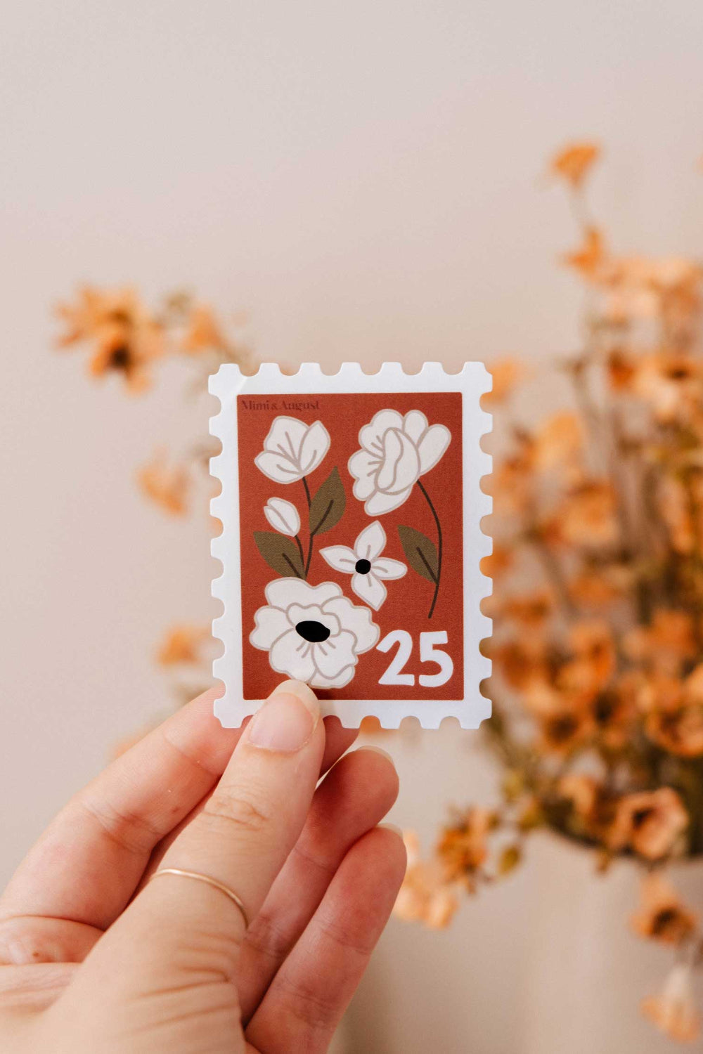 Hand holding a decorative flower stamp sticker and number 25 from Mimi and August.