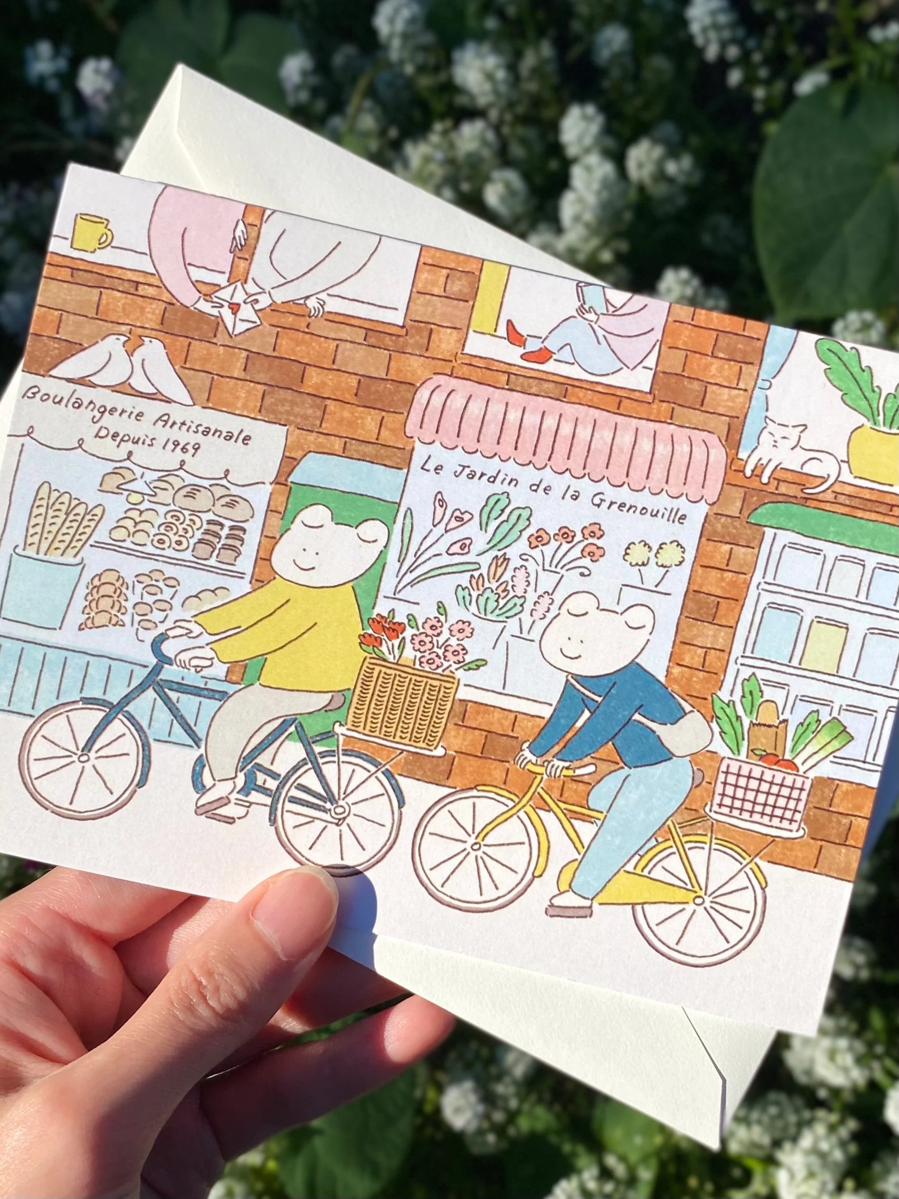 Illustrated greeting card with two frogs on bicycles held by a hand in the city