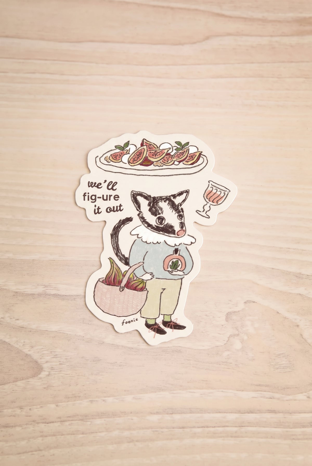 A die-cut sticker featuring an illustrated badger character holding a plate of figue and a drink, with the text 'we'll fig-ure it out', placed on a light wood surface. illustrated by Foonie at la maison garçonne in Montreal.