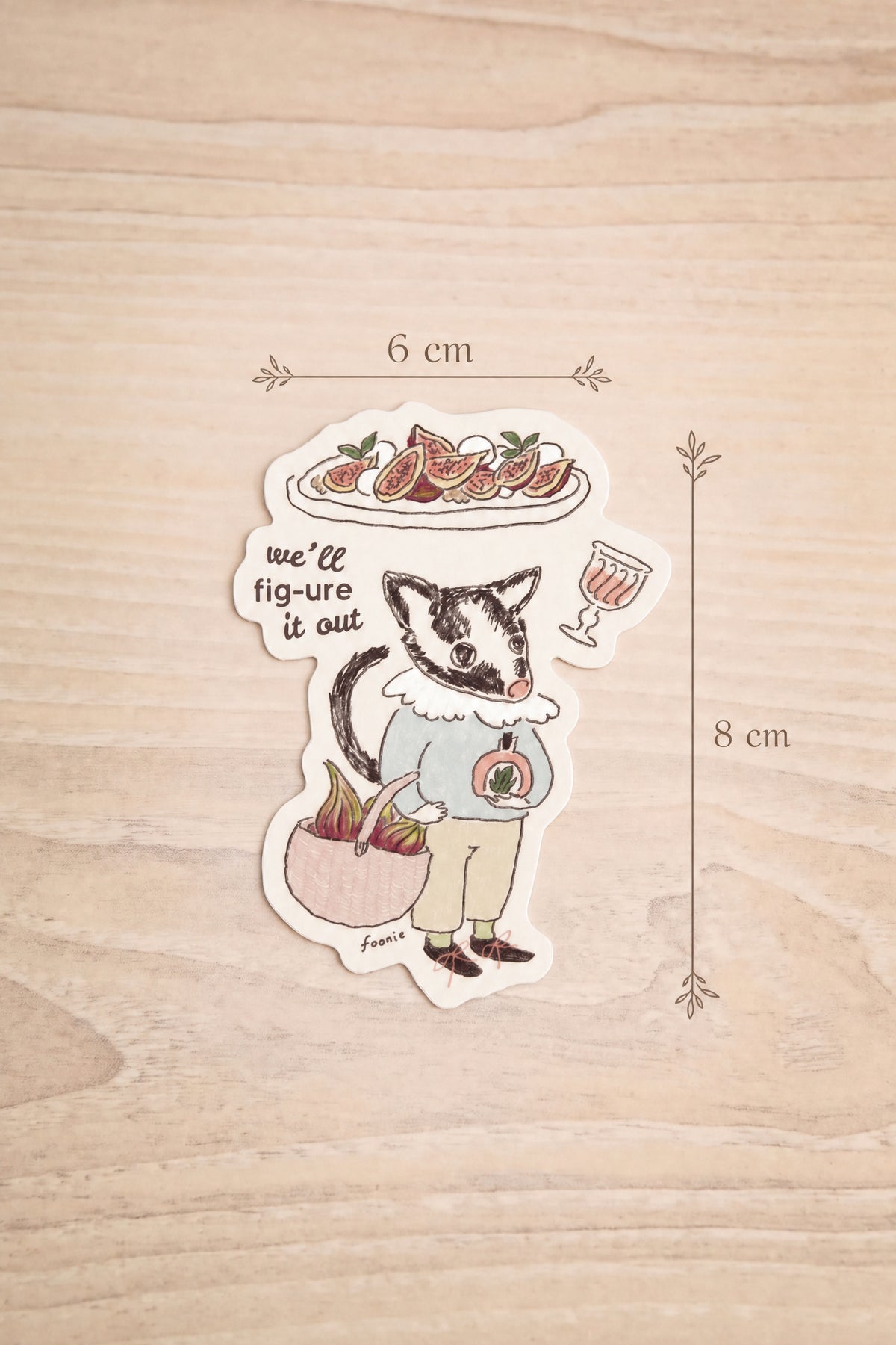 Dimension 6cm x 8cm of a a die-cut sticker featuring an illustrated badger character holding a plate of figue and a drink, with the text 'we'll fig-ure it out', placed on a light wood surface. illustrated by Foonie at la maison garçonne in Montreal.