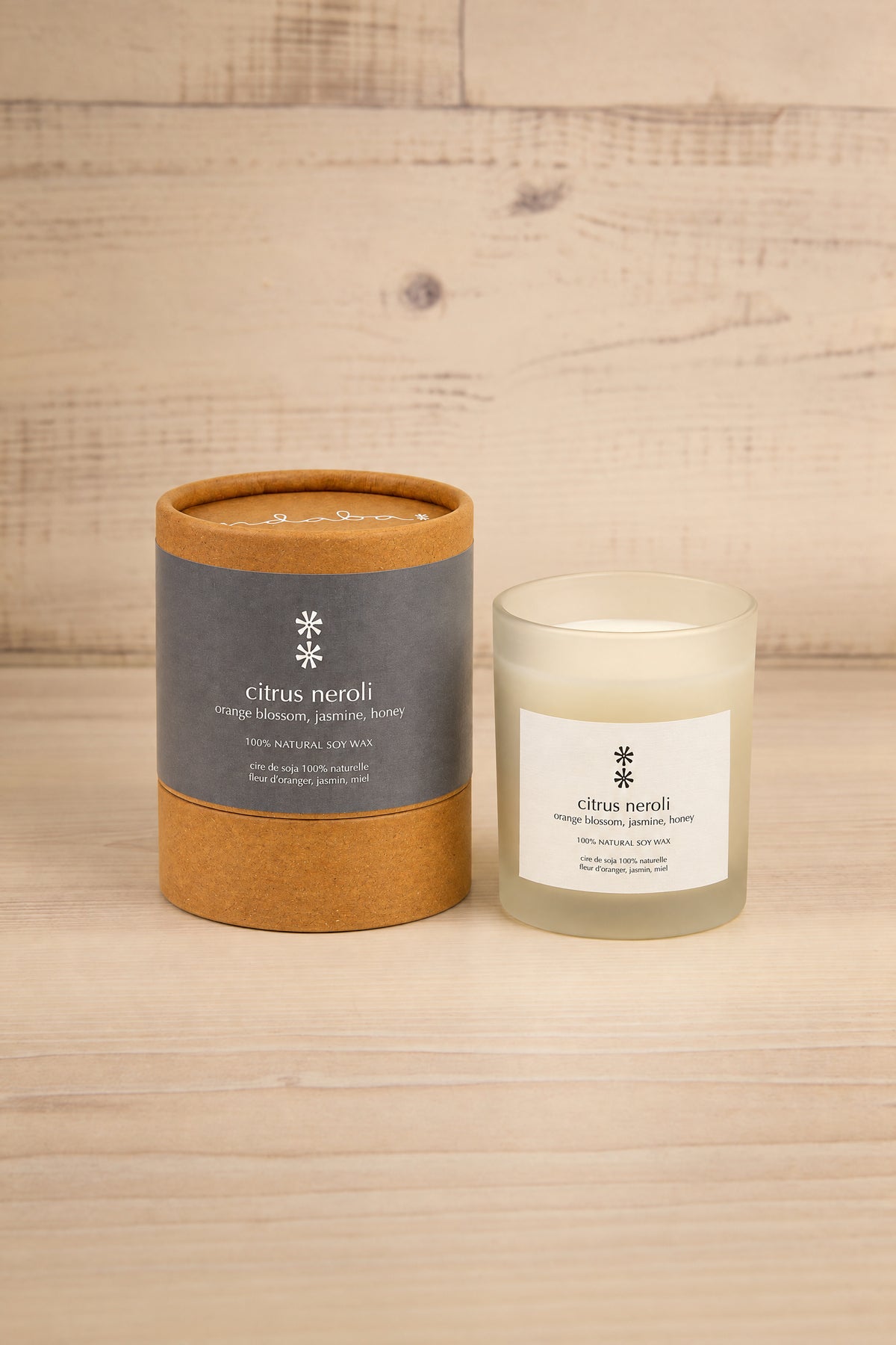 A candle product shown with its round kraft paper packaging tube and the frosted glass candle jar beside it, set on a light wood surface against a light wood plank background. The labels read citrus neroli at la maison garçonne.