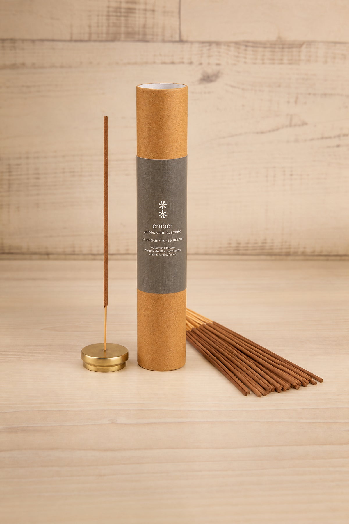 A tube of incense sticks stands upright on a light wood surface with a brass incense holder and several incense sticks beside it. The tube is kraft brown with a dark gray label reading 'EMBER' at la maison garçonne in montreal.