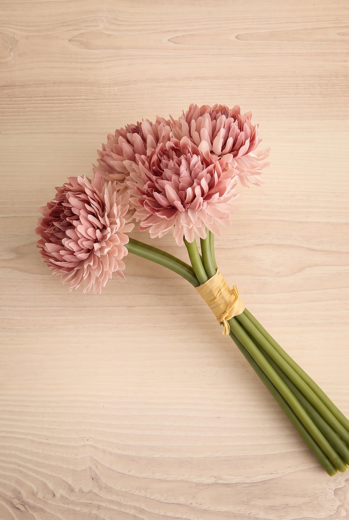 A small bouquet of pink chrysanthemum-like artificial flowers with long green stems, tied together with a natural tan band, placed diagonal on a light wood surface at la maison garçonne in montreal.